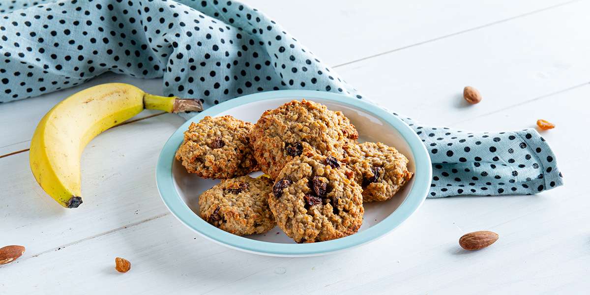 Goldbraune Rosinen Hafer Cookies in einer Schüssel. Daneben eine frische Banane, Mandeln und Rosinen.