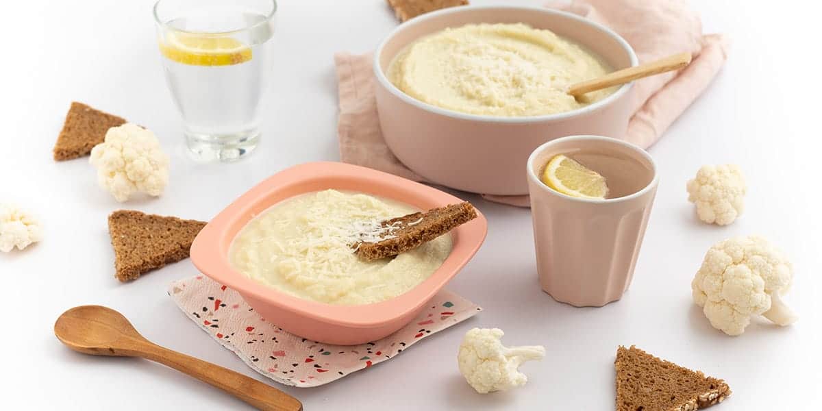 Blumenkohl Suppe in einer Schüssel serviert. Ein Brotstick steckt in der Suppe. Daneben steht ein Becher mit Wasser und dahinter eine große Schüssel mit der Suppe