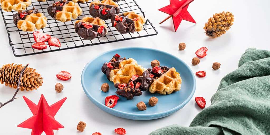 Waffelkekse auf einem Teller serviert. Dahinter sind weitere Waffelkekse auf einem Gitter. Weihnachtliche Deko mit roten Sternen und Tannenzapfen.