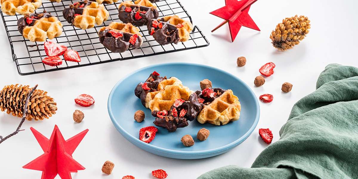 Waffelkekse auf einem Teller serviert. Dahinter sind weitere Waffelkekse auf einem Gitter. Weihnachtliche Deko mit roten Sternen und Tannenzapfen.