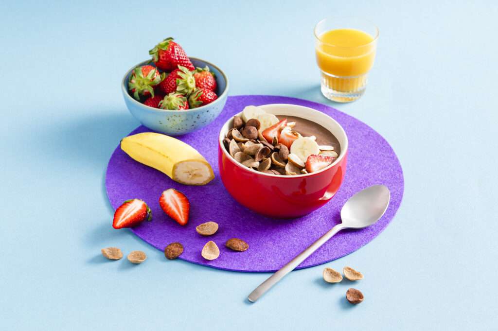 Schoko Bowl in einer Schüssel angerichtet. Daneben steht ein Glas Orangensaft und liegt ein Löffel. Frische Erdbeeren und ein Stück Banane liegen zur Deko dabei.