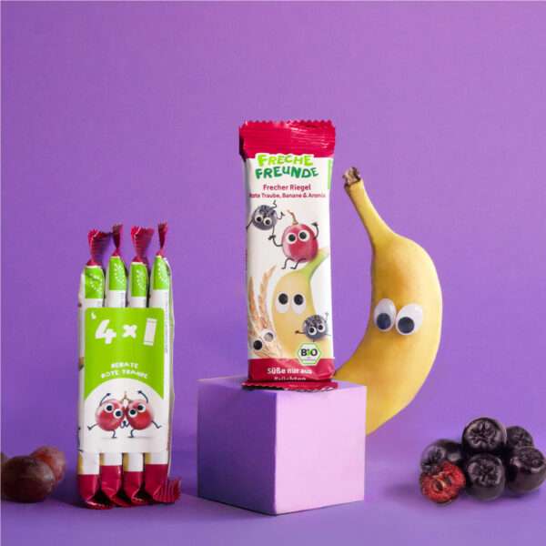 Die Verpackungseinheit mit 4 Freche Freunde Bio Riegeln Rote Traube, Banane & Aronia steht neben einem einzeln verpackten Riegel. Links und rechts posieren die frechen Charaktere mit Kulleraugen vor lilafarbenem Hintergrund – begleitet von echten Früchten wie Banane, Traube und Aroniabeeren.