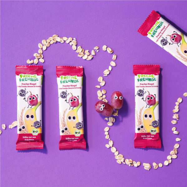Vier Freche Freunde Bio Riegel Rote Traube, Banane & Aronia liegen nebeneinander auf einem lilafarbenen Untergrund. Eine Spur aus Haferflocken schlängelt sich über das Bild. Zwei fröhliche rote Trauben mit Kulleraugen ergänzen die Szene und machen das Bild besonders frech.