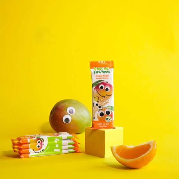 Freche Freunde Bio Riegel Mango & Orange vor leuchtend gelbem Hintergrund – mit echter Mango, Orangenspalte und frechen Kulleraugen. Momo Mango grinst neben dem aufrecht stehenden Riegel.