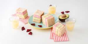 Gemüse-Sandwich mit rosa, grünem & beigem Aufstrich auf einem Teller, daneben Rote Bete Stückchen und aufgeschnittene Avocado