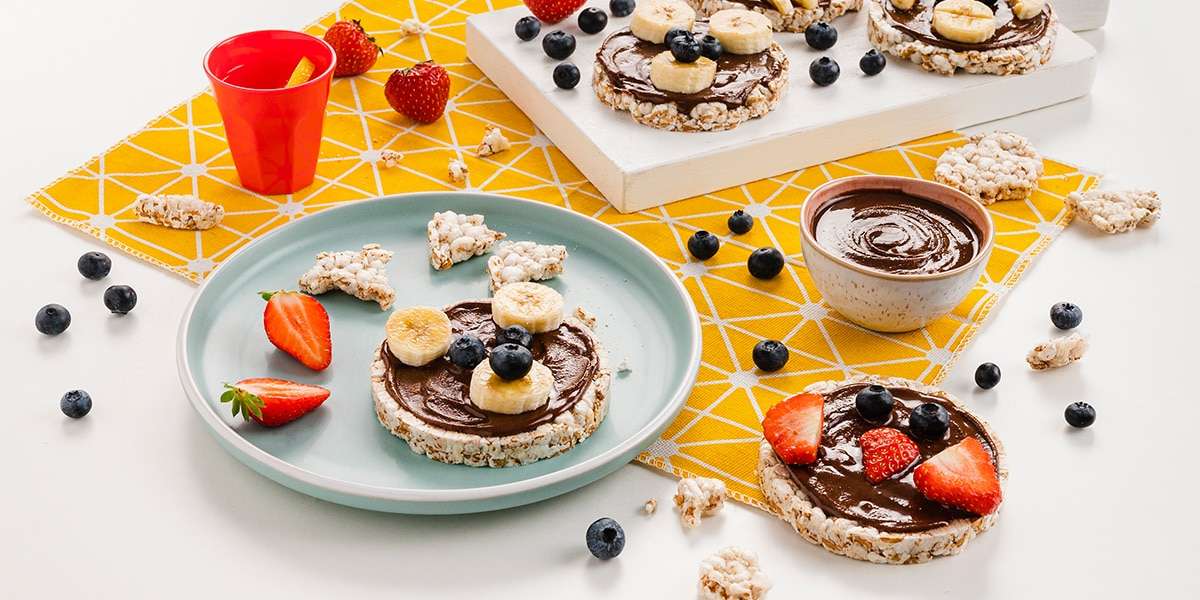 Reiswaffeln mit Haselnuss-Kakao Creme bestrichen und mit Gesichtern aus Bananenscheiben, Blaubeeren und Erdbeeren dekoriert.