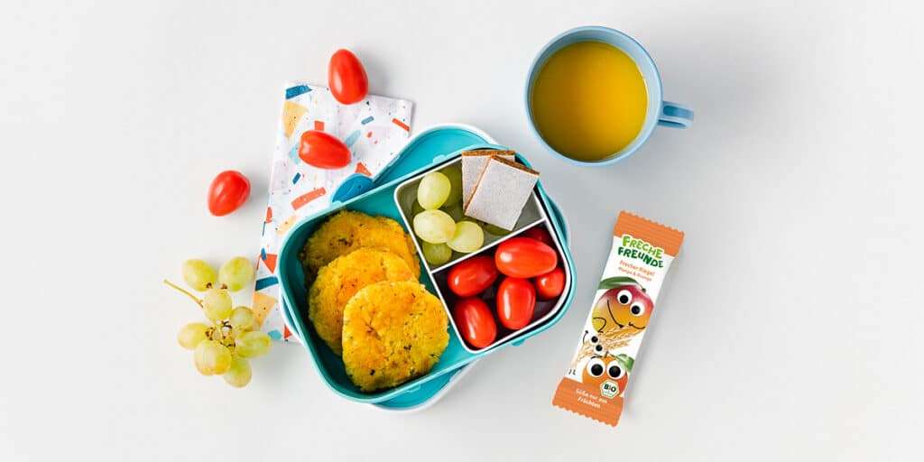 Lunchbox mit Hirsebratlingen, Tomaten, Weintrauben und Freche Freunde Mango-Orange Riegel gefüllt.