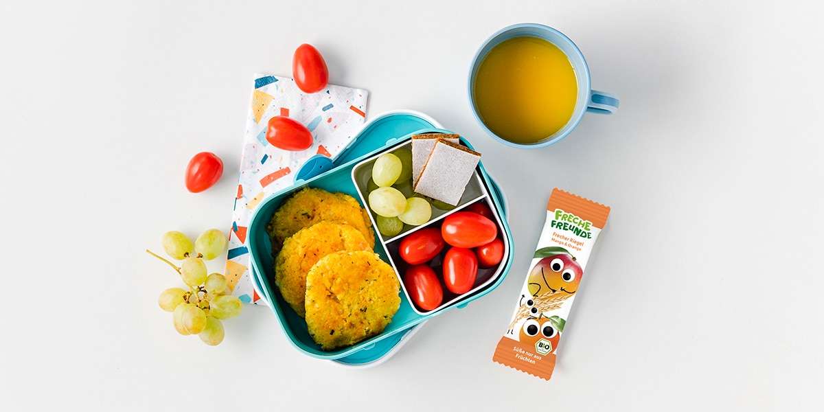 Lunchbox mit Hirsebratlingen, Tomaten, Weintrauben und Freche Freunde Mango-Orange Riegel gefüllt.