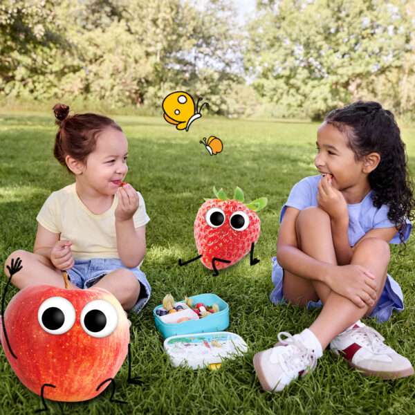 Zwei lachende Kinder sitzen im Gras beim Picknick – zwischen ihnen eine geöffnete Freche-Freunde-Brotdose mit fruchtigen Snacks. Edda Erdbeere und Alfred Apfel sorgen mit frechen Grimassen für gute Laune.