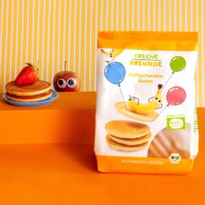 Pancakes Banane Tüte auf orangenem Hintergrund, mit Pancakes und Erdbeere, sowie Alfred Apfel mit Wackelaugen