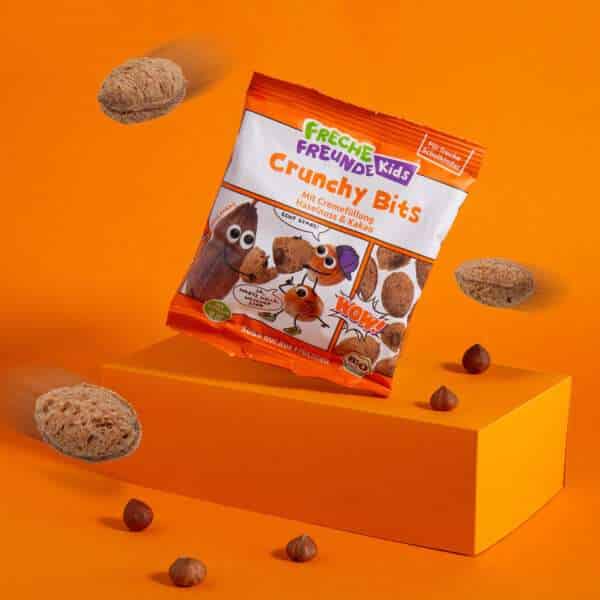 Freche Freunde Kids Bio Crunchy Bits auf einem orangem Podest und Hintergrund und rundherum fliegende Crunchy Bits.