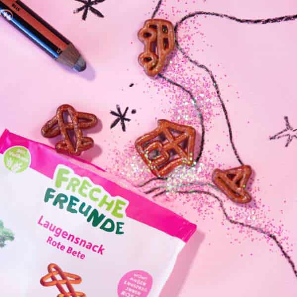 Freche Freunde Bio Laugensnack Rote Bete – Brezeln in Auto-, Flugzeug-, Haus- und Bootform liegen auf einer rosa Glitzerstraße mit gezeichneten Sternen. Die Verpackung schaut frech ins Bild.