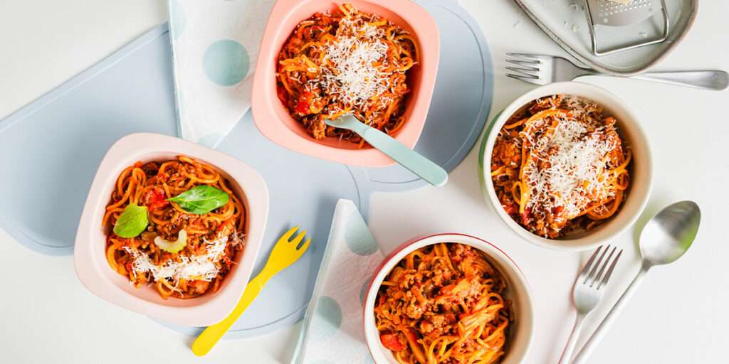 One Pot Spaghetti Bolognese in 4 Tellern angerichtet und mit Parmesan dekoriert. Links und rechts liegen Messer und Gabel sowie Kinderbesteck.