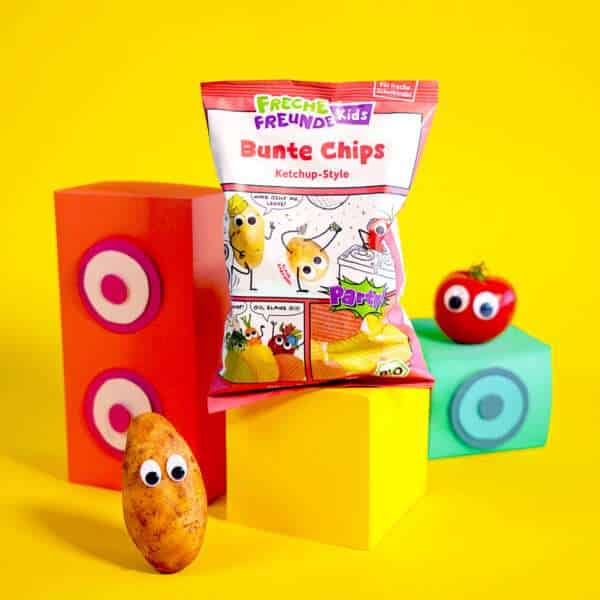 Freche Freunde Kids Bunte Chips Ketchup-Style Verpackung im Comic Design auf gelben Podest und Hintegrund, daneben Klaus Kartoffel und Tom Tomate mit Wackelaugen.