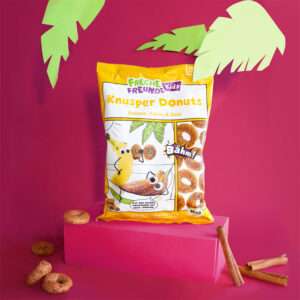 Freche Freunde Bio Knusper Donuts Banane, Kakao & Zimt Verpackung im Comicdesign auf einem rosa Podest umgeben von Zimtstangen und kleinen Donuts.