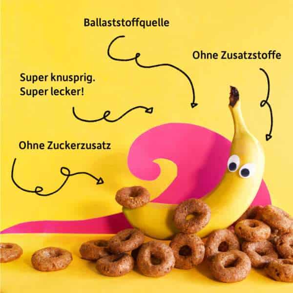 Freche Freunde Bio Knusper Donuts mit Banane, Kakao & Zimt tummeln sich um Bob Banane mit Wackelaugen auf buntem Hintergrund. Aussagen wie "Ballaststoffquelle", "Ohne Zusatzstoffe" und "Ohne Zuckerzusatz"..
