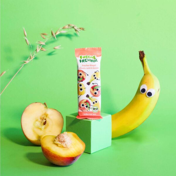 Der Freche Freunde Riegel Pfirsich, Apfel & Banane steht auf grünem Hintergrund neben frischen Früchten und Getreide. Neben dem Riegel liegt eine Banane mit frechen Augen – das ist Bob Banane.