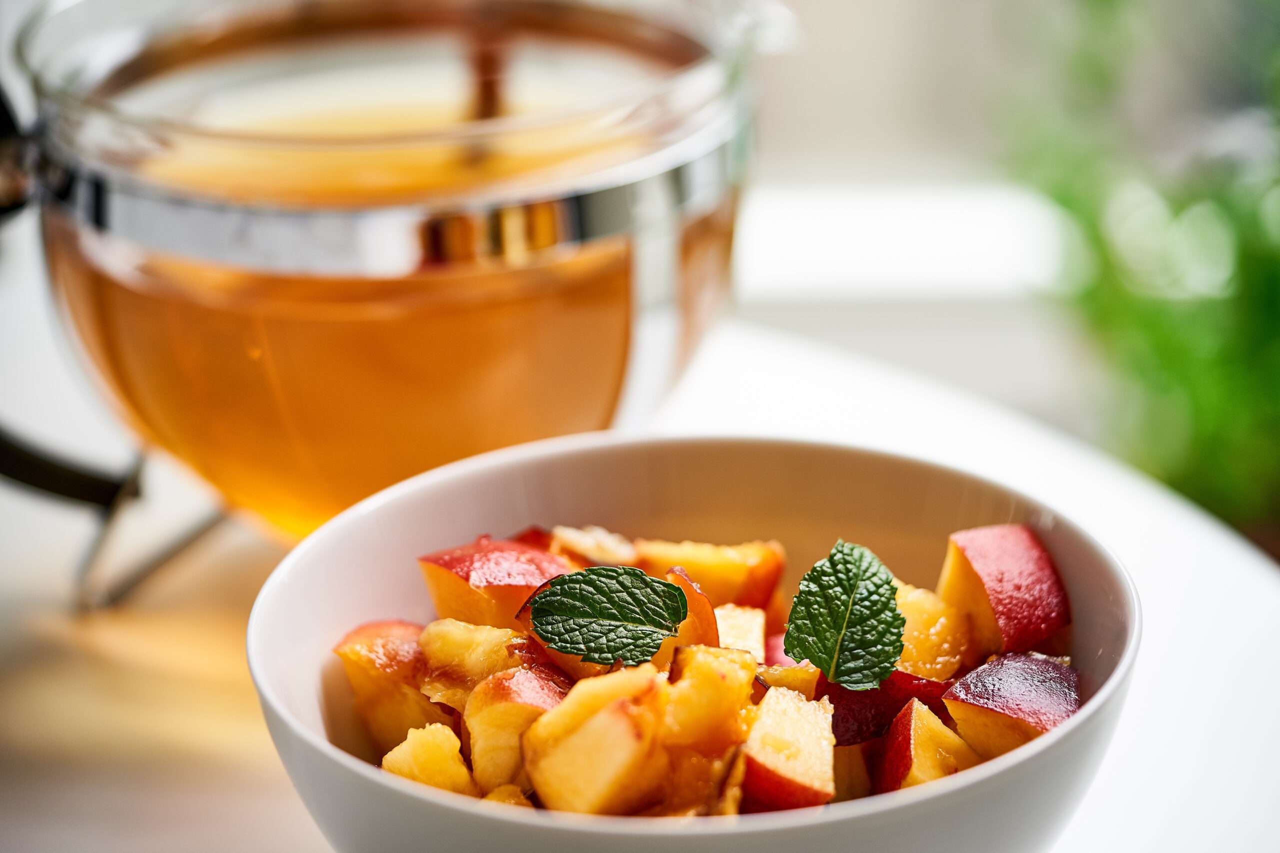 Auf einem Tisch steht eine Schüssel mit Pfirsich und Minze, dahinter eine Teekanne.