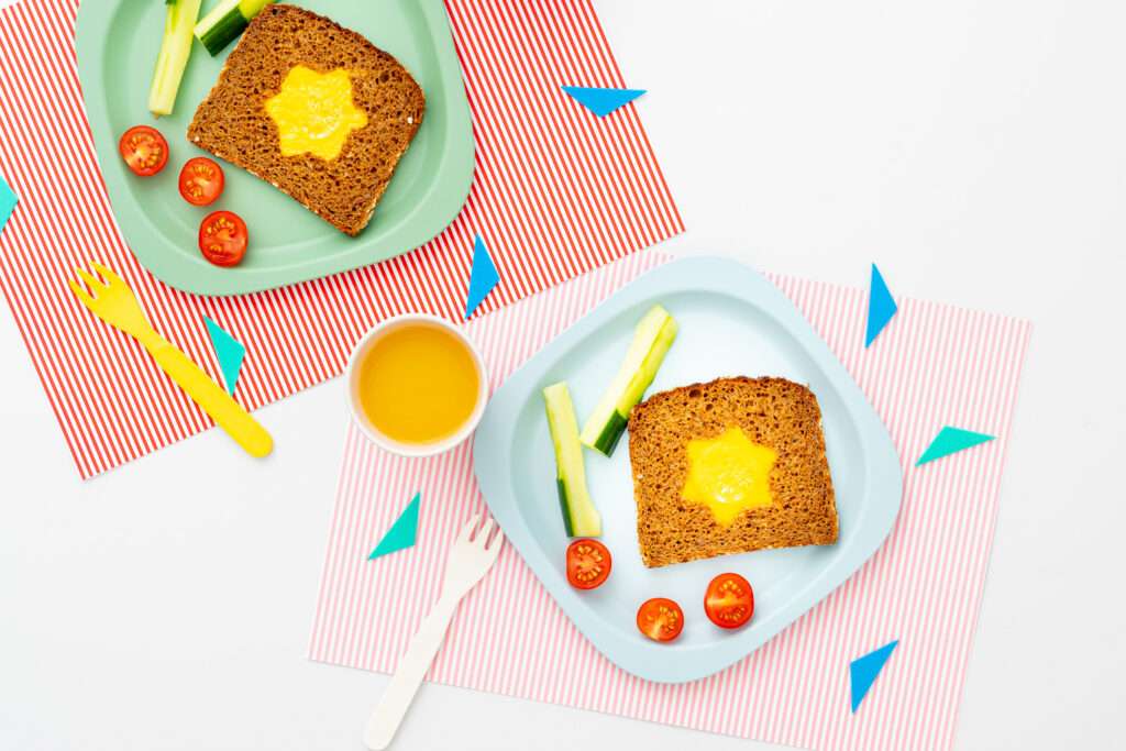 Sternförmies Ei im Brot auf einem Teller serviert mit Gurkensticks & Tomaten dazu ein Becher Orangensaft.