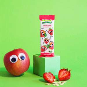 Freche Freunde Bio Frecher Riegel Apfel & Erdbeere posiert auf grünem Podest. Neben dem Riegel mit Alfred Apfel und Edda Erdbeere eine echte Erdbeere und ein Apfel mit Wackelaugen.