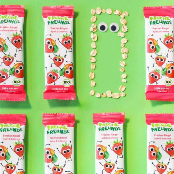 Freche Freunde Bio Frecher Riegel Apfel & Erdbeere umgeben von Haferflocken, die sich zu einem Riegel mit Augen formieren. Ein freches Arrangement, das zeigt, was alles in dem fruchtigen Riegel steckt.