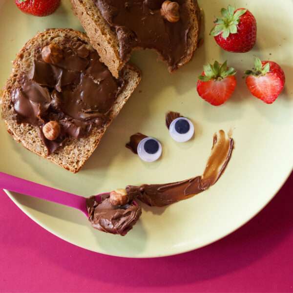 Lustig angerichteter Teller mit zwei Scheiben Brot mit Freche Freunde Kids Bio Creamy Haselnuss-Kakao-Creme, frischen Erdbeeren und einem frechen Schoko-Gesicht aus Creme, Wackelaugen und einem Löffel mit Hassan Haselnuss.