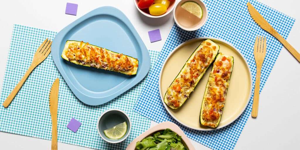 Gefüllte Zucchini überbacken mit Käse auf Tellern serviert. Dazu steht ein kleiner Salat, Trinkgläser und Besteck.
