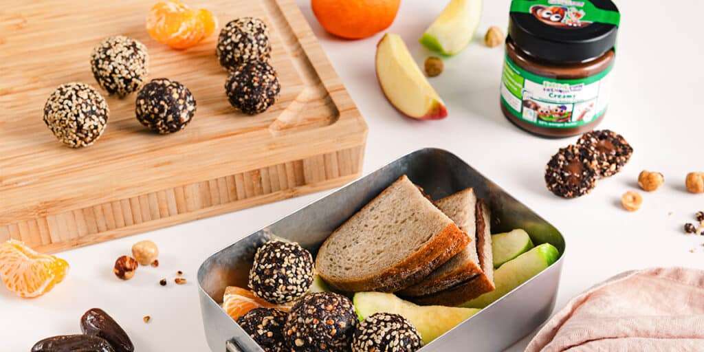Bliss Balls liegen auf einem Brett, daneben steht eine Lunchbox mit Bliss Balls und einem Brot mit Haselnuss-Kakao-Creme bestrichen. Im Hintergrund steht ein Glas Freche Freunde Creamy Haselnuss-Kakao-Creme, Haselnüsse und Apfelspalten.