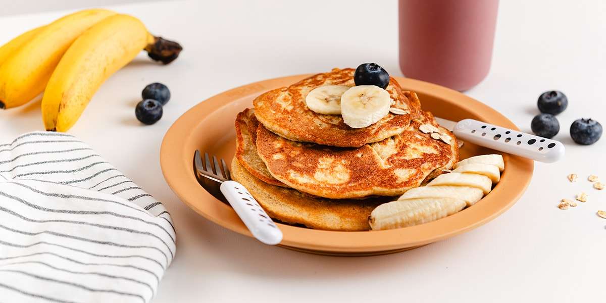 Bananenpfannkuchen auf einem Teller serviert mit frischen Bananenscheiben und Baubeeren. Im Hintergrund liegen frische Bananen und Blaubeeren. Hinter dem Teller steht ein Becher mit Wasser.