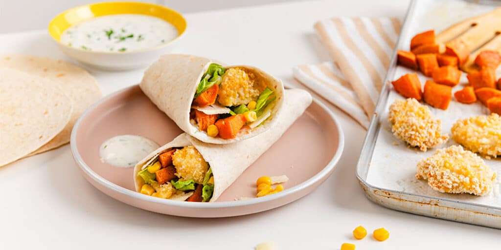 Zwei leckere Crispy Nuggets Wraps serviert auf einem Teller. Daneben liegt das Blech mit Chicken Nuggets und Süßkartoffeln aus dem Ofen und eine eine Schüssel mit einen Dip.