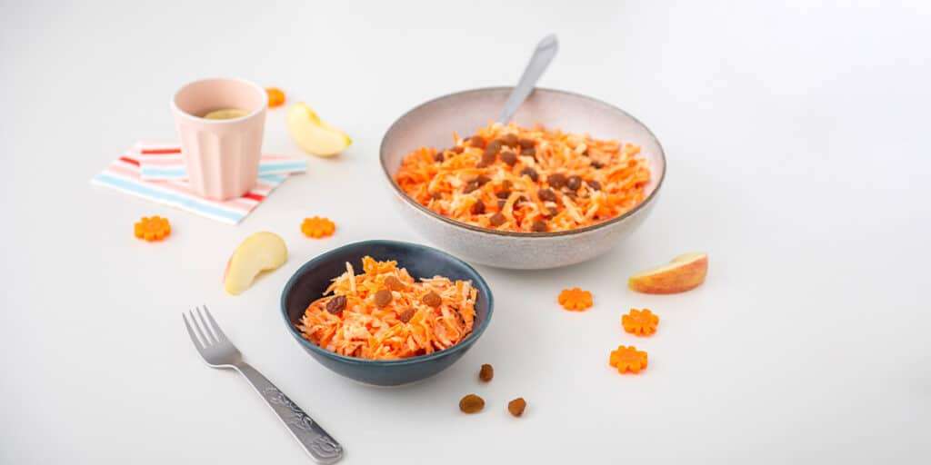 Karotten Apfel Salat in einer Schüssel serviert, daneben steht eine kleine Schüssel mit Salat & einer Gabel sowie ein Becher mit Wasser