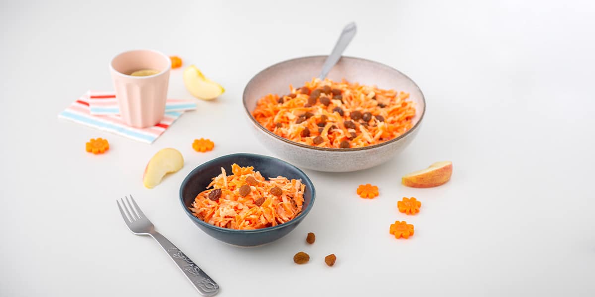 Karotten Apfel Salat in einer Schüssel serviert, daneben steht eine kleine Schüssel mit Salat & einer Gabel sowie ein Becher mit Wasser