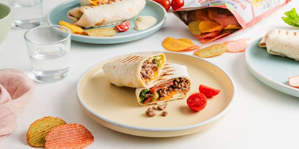 Cheeseburger Wraps auf Tellern serviert. Dazu liegen Freche Freunde Bunte Chips und frische Tomate sowie ein Glas Wasser.