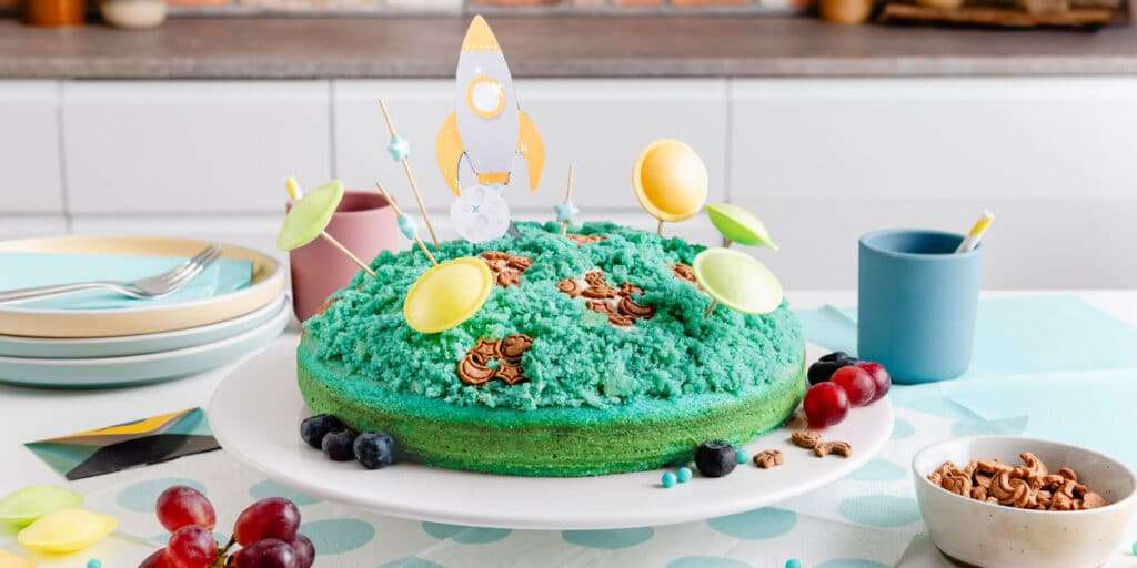 Weltraumkuchen auf einem Teller serviert und mit Weltraum Deko dekoriert. Daneben stehen Teller und ein Becher.