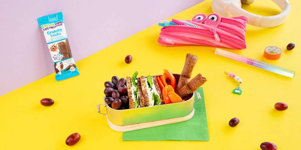 Ei Sandwich in einer Brotdose zusammen mit Trauben, Paprikastreifen und Freche Freunde Kids Crunchy Sticks. Daneben liegt eine Federtasche, Lineal und Kopfhörer.