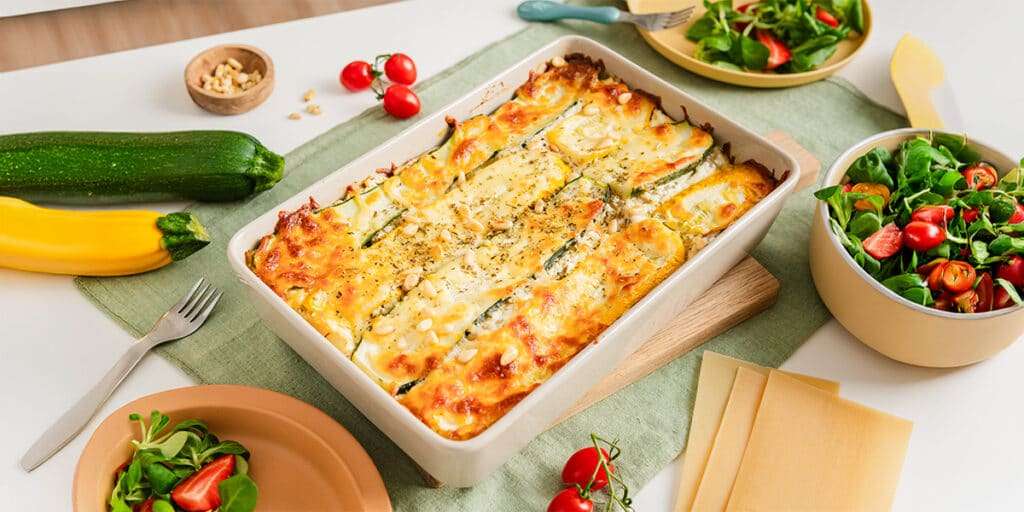 Zucchini Lasagne in einer Auflaufform auf einem Tisch angerichtet. Dazu steht eine Schüssel mit frischem Salat und Teller.